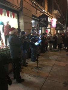 A group of musicians play holiday music in front of stores decorated for Christmas. 